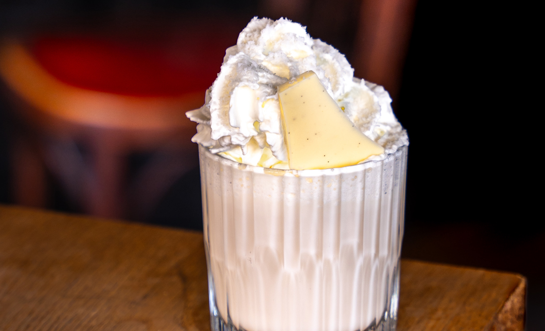 A small glass is placed at the edge of a wooden table. The glass contains a hot chocolate topped with whipped cream and white chocolate. 