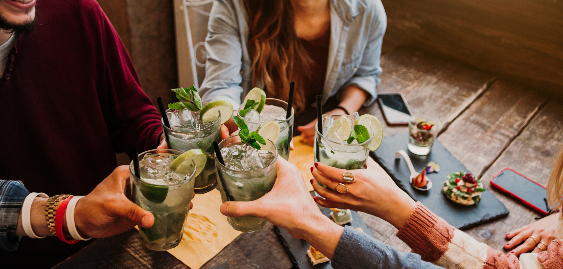 A group of people are sat at a wooden table. They are holding up their drinks, all of which have mint leaves. 