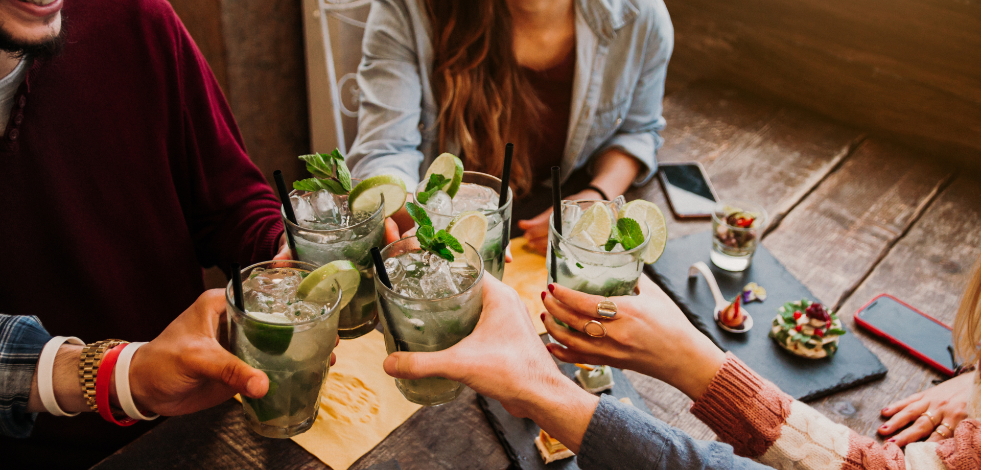A group of people are sat at a wooden table. They are holding up their drinks, all of which have mint leaves.