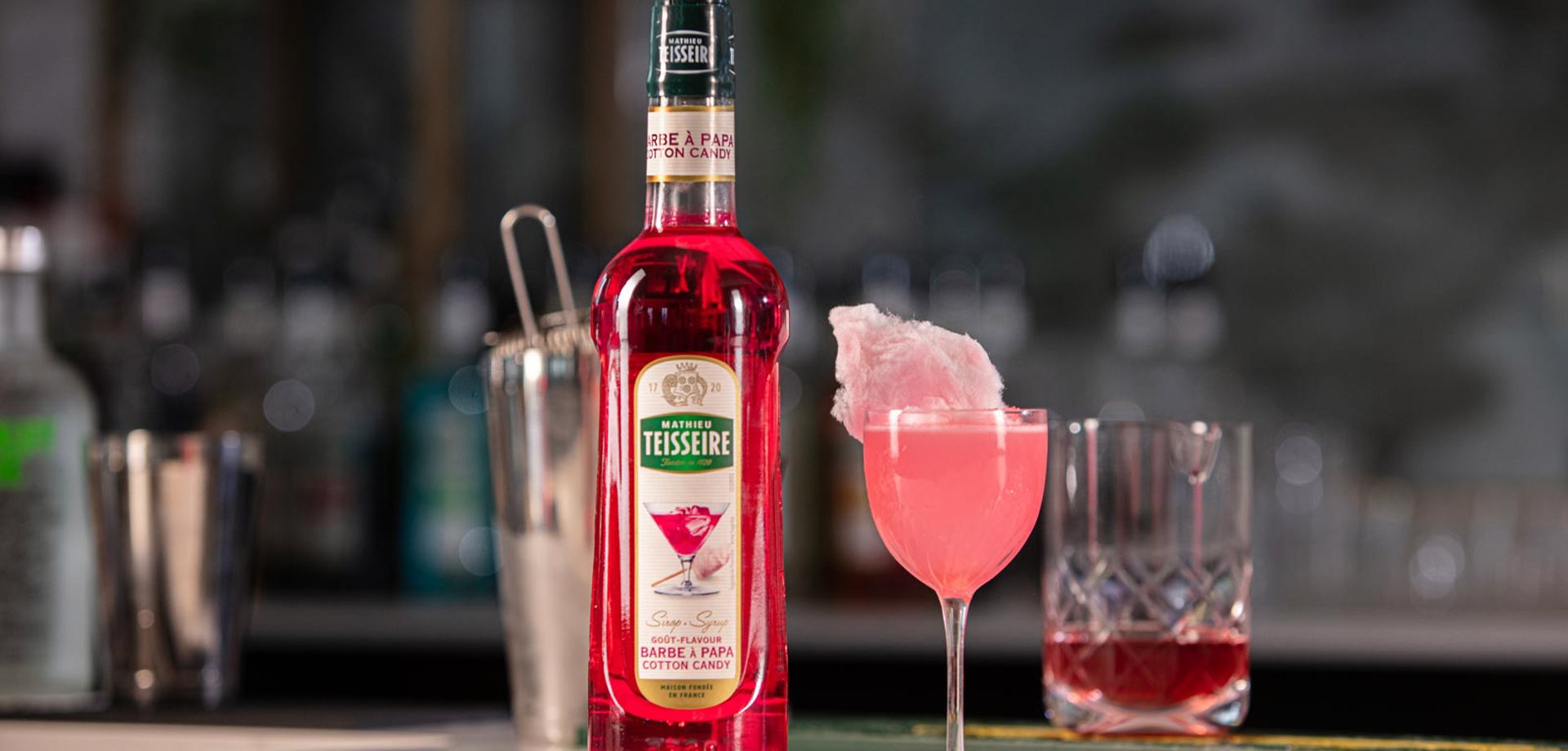 On a bar surface there is a bottle of Mathieu Teisseire cotton candy syrup, next to a glass filled with a pink drink and topped with cotton candy.