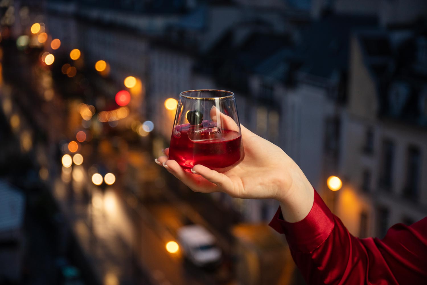 A hand holds a red drink over a scenic view. In the background it is night, and the street is illuminated by car headlights and streetlights.