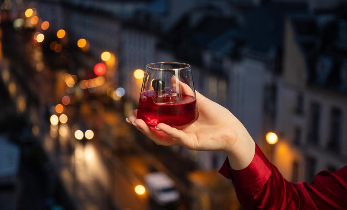 A hand holds a red drink over a scenic view. In the background it is night, and the street is illuminated by car headlights and streetlights.