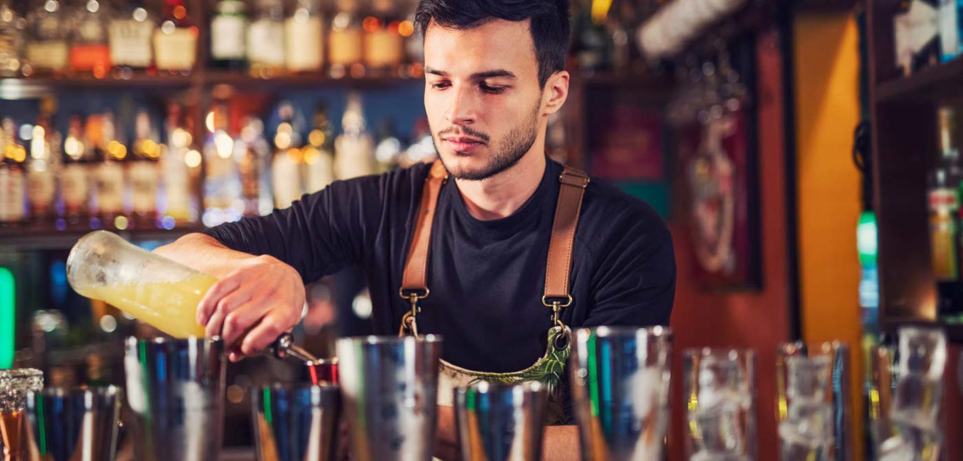 A male bartender standing behind a bar preparing a cocktail. He is pouring a spirit into a shot glass. On the bar there are different size cocktail shakers.