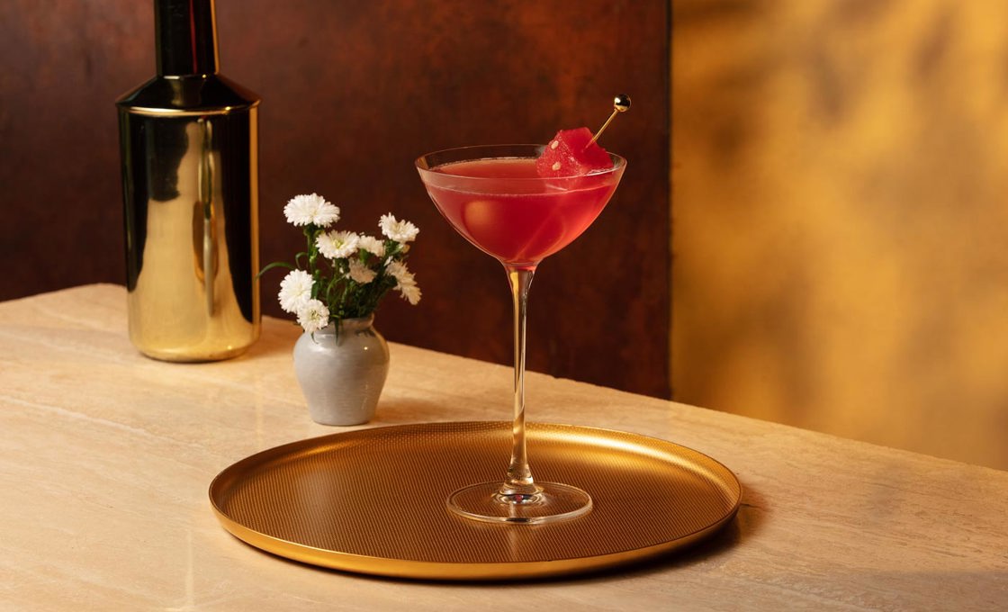 Atop a gold tray is a coupe glass filled with a red liquid, and a golden cocktail stick with a cube of watermelon. In the background is a small vase of flowers and a golden cocktail shaker.