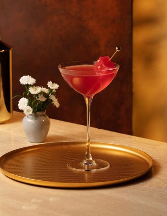 Atop a gold tray is a coupe glass filled with a red liquid, and a golden cocktail stick with a cube of watermelon. In the background is a small vase of flowers and a golden cocktail shaker.