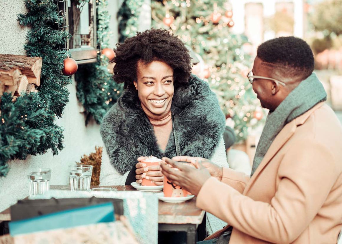 A man and a woman in winter coats sitting outside a cafe. They are laughing and talking whilst drinking cups of hot chocolate. 