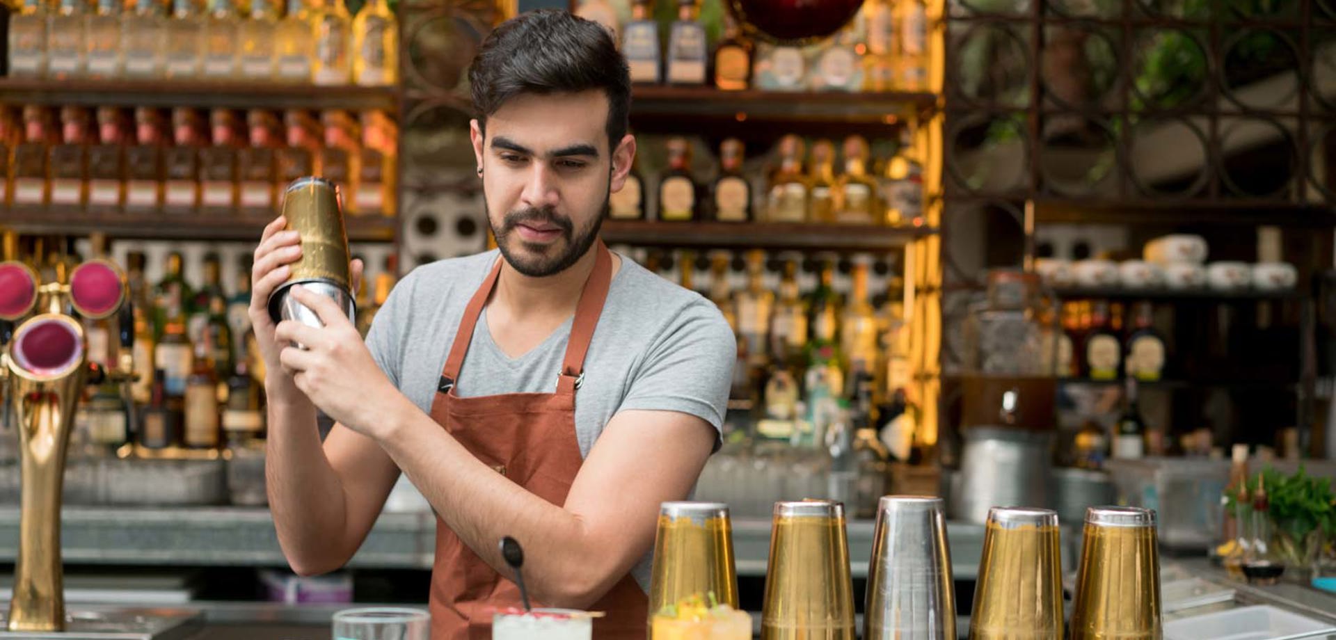 A male bartender standing behind a bar shaking a cocktail in a shaker. The bar is white marble and has glasses and cocktail shakers on the surface.