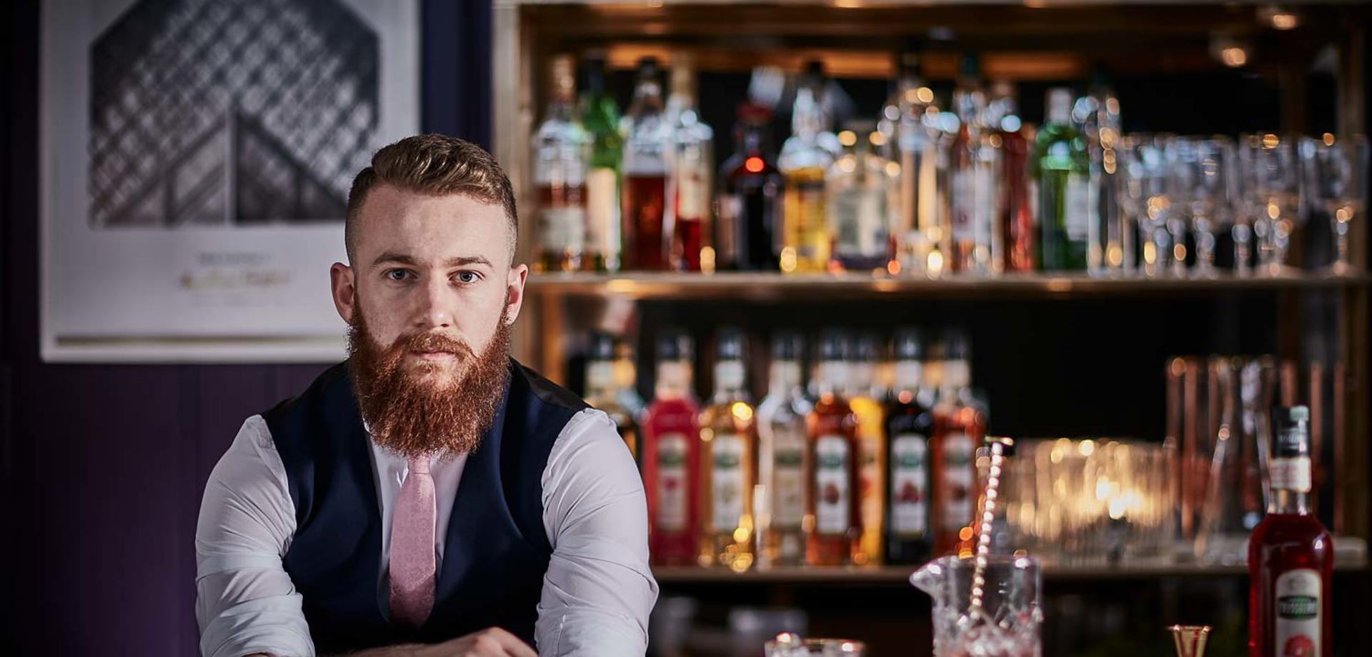 A male bartender standing at a bar. The backbar is filled with glasses, Mathieu Teisseire syrup bottles and cocktail shakers. There is a cocktail mixing glass on the bar filled with with red liquid and a bar spoon.