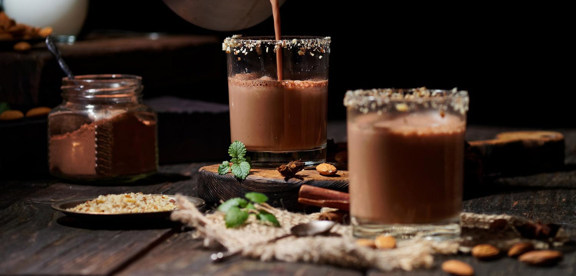 Two rocks glasses on a table filled with hot chocolate and garnished with crushed nuts around the rim of the glass. There is a hand out of shot pouring the chocolate into one of the glasses from a pan.