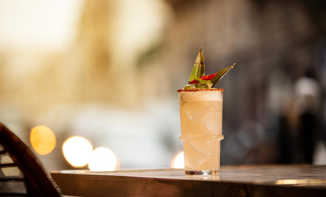 A clear glass contains a white drink topped with pineapple leaves. It sits on a long outdoor table and in the background we can see a long street lit by bright light.