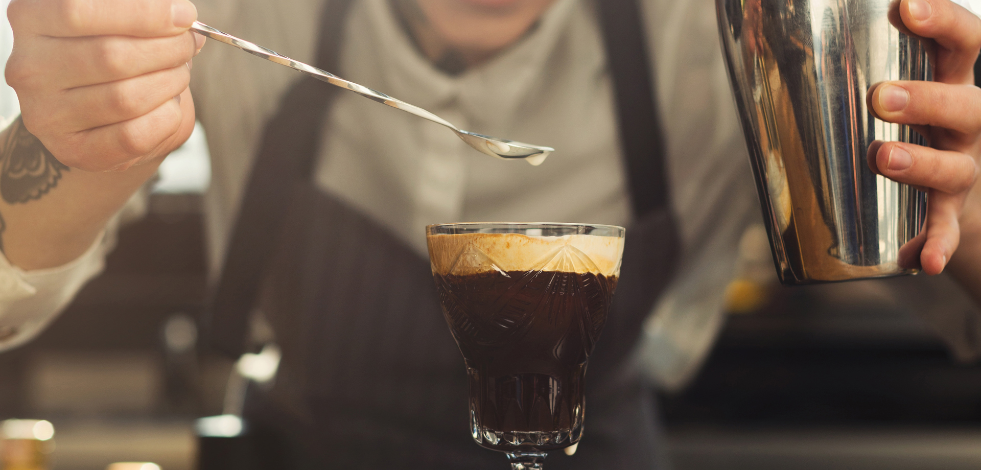 A woman wearing an apron stands behind a counter, she is holding a cocktail shaker and a bar spoon. She is adding the finishing touches to a coffee based drink. 