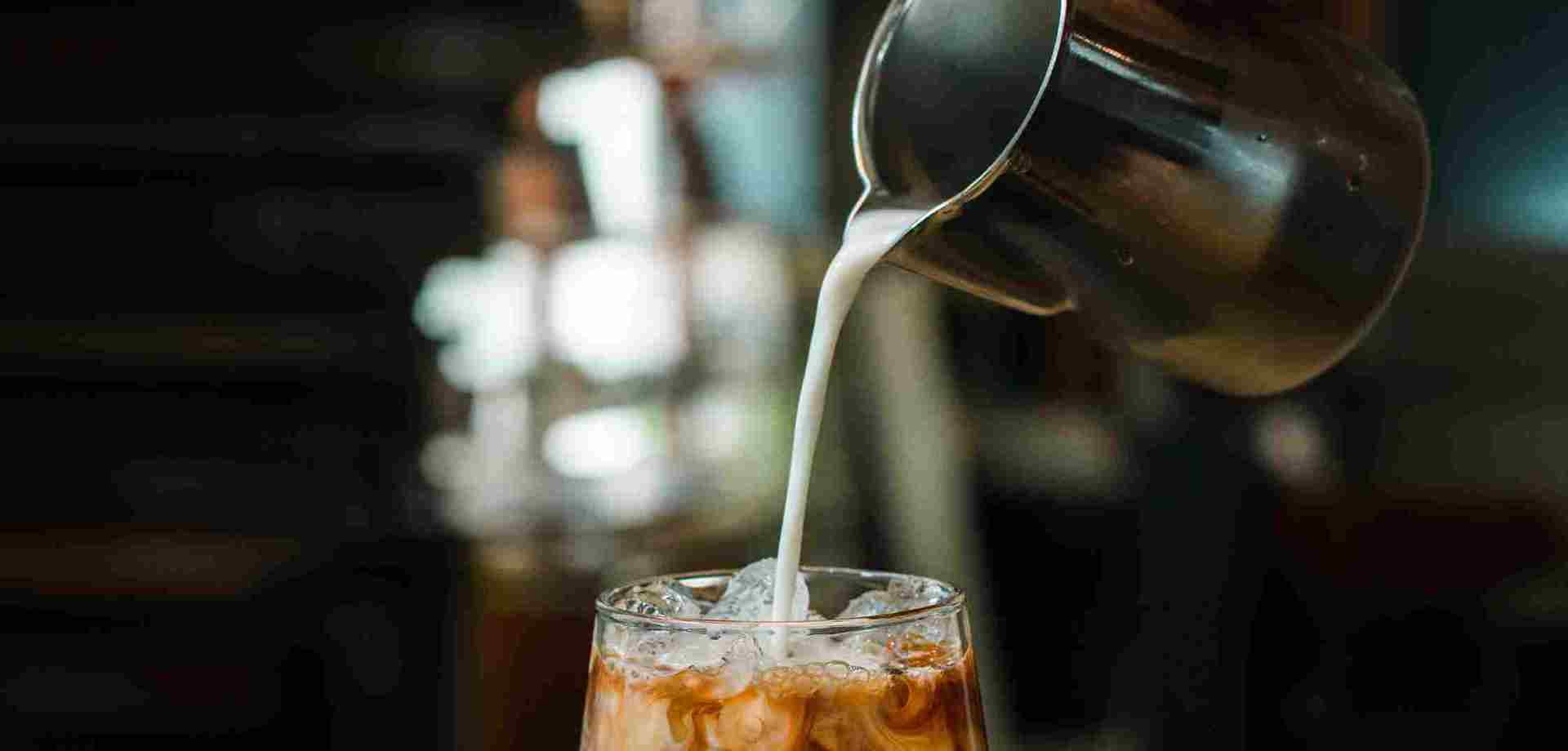 A glass of iced coffee sits on a black counter. Milk is being poured into the coffee from a metal jug.