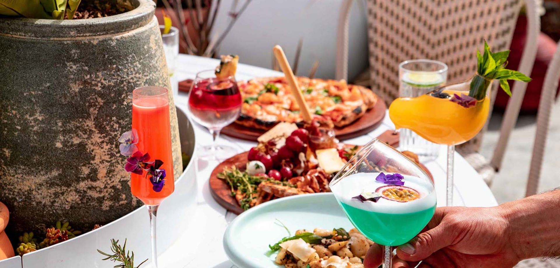 A table with a selection of food and cocktails. The cocktails are in martini glasses and flutes and are brightly coloured with beautiful floral garnishes. 
