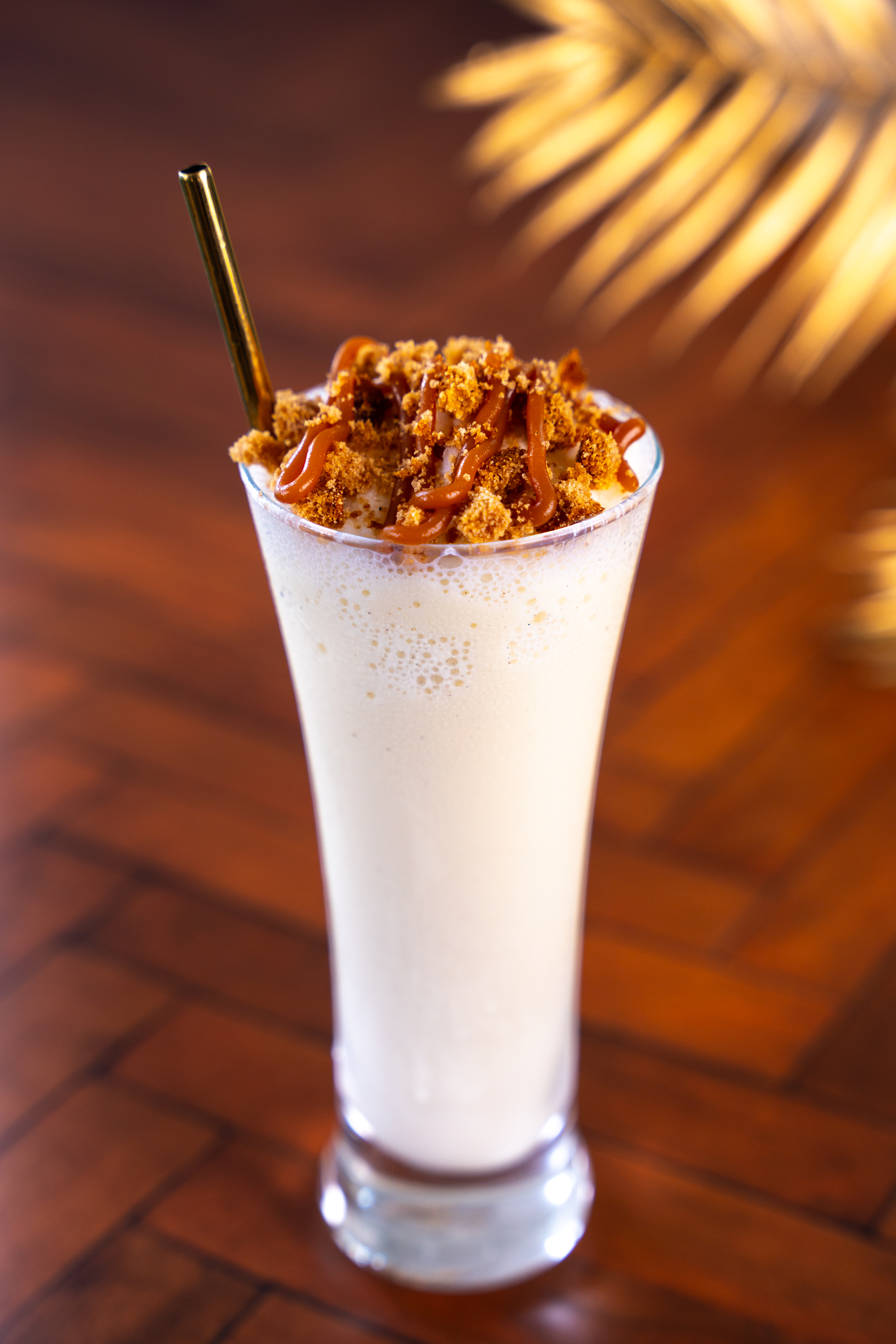 On a polished wooden surface there is a tall milkshake glass, containing a white coloured milkshake. The drink is topped with gingerbread crumbs and caramel sauce. 