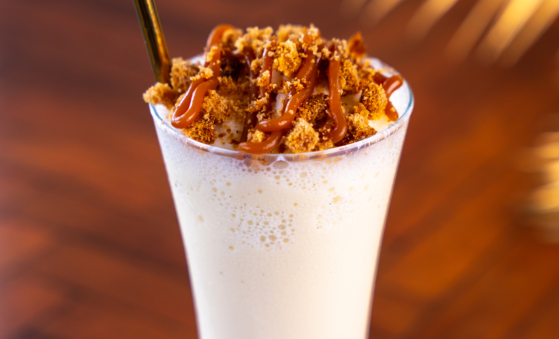 On a polished wooden surface there is a tall milkshake glass, containing a white coloured milkshake. The drink is topped with gingerbread crumbs and caramel sauce. 