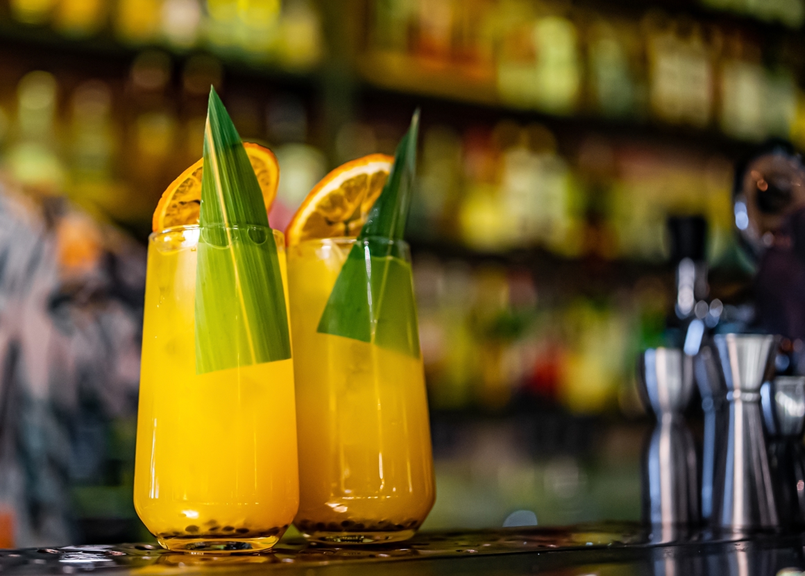 Two cocktails are placed on a bar. They are both in tall glasses, and contain orange coloured drinks. They are topped with orange slices and green leaves.