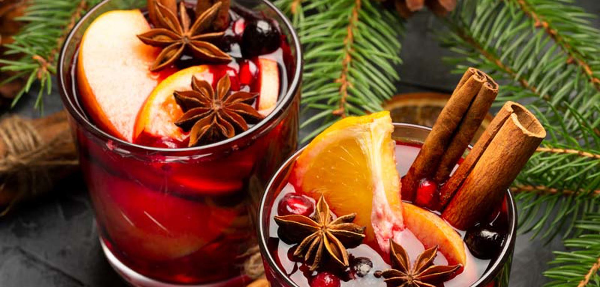 Two rocks glasses on a table filled with a red winter cocktail and garnished with slices of orange, apple and cinnamon sticks. There are pine cones and fir tree brances scattered around the glasses.