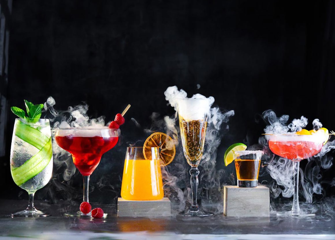 Six cocktails lined up on a grey surface. Each cocktail is in a different glass, with different liquids inside. Each glass is smoking with dry ice.