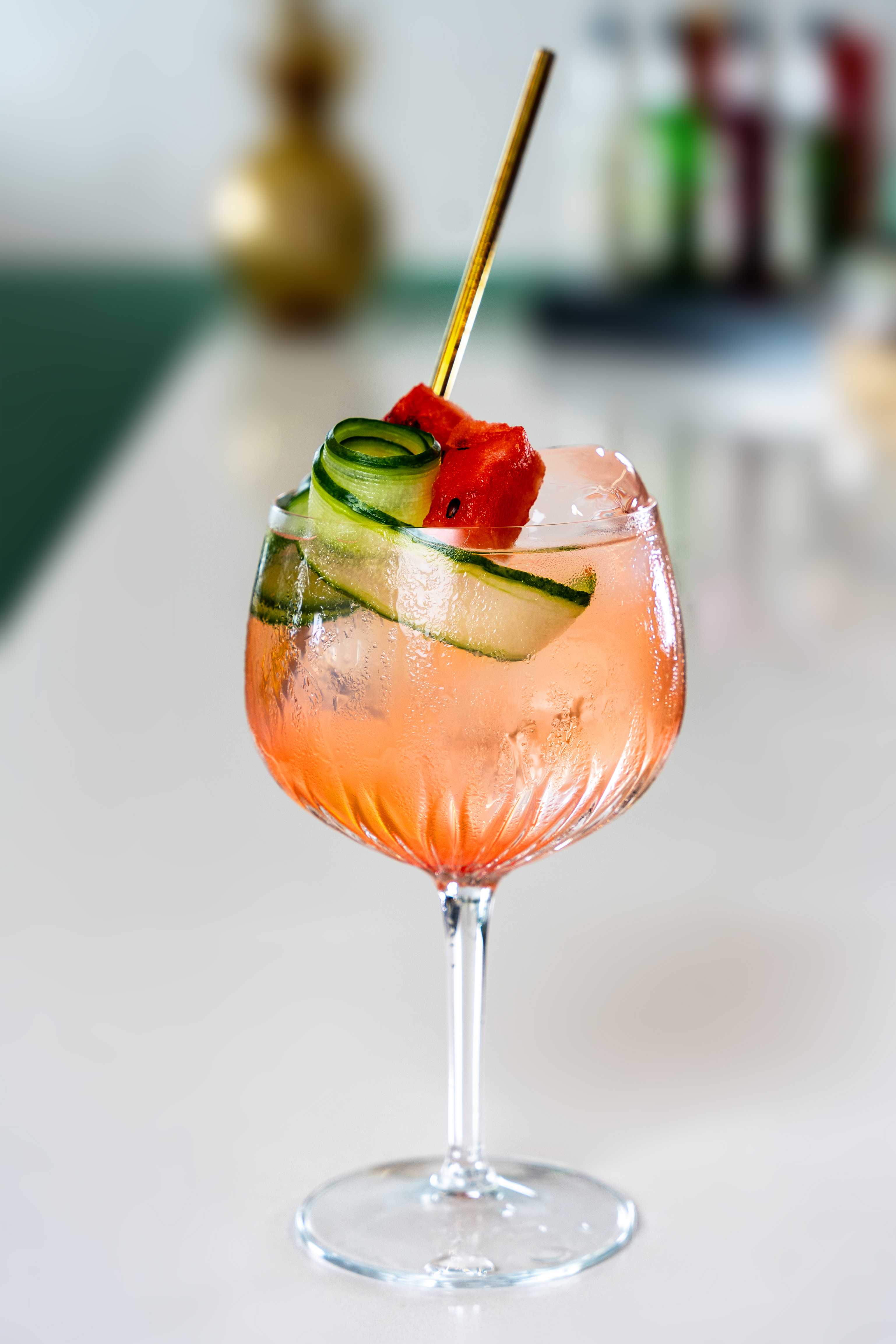 A large copa glass is placed on a marble bar top. The glass contains a pale pink drink, and is topped with a ring of fresh cucumber. 