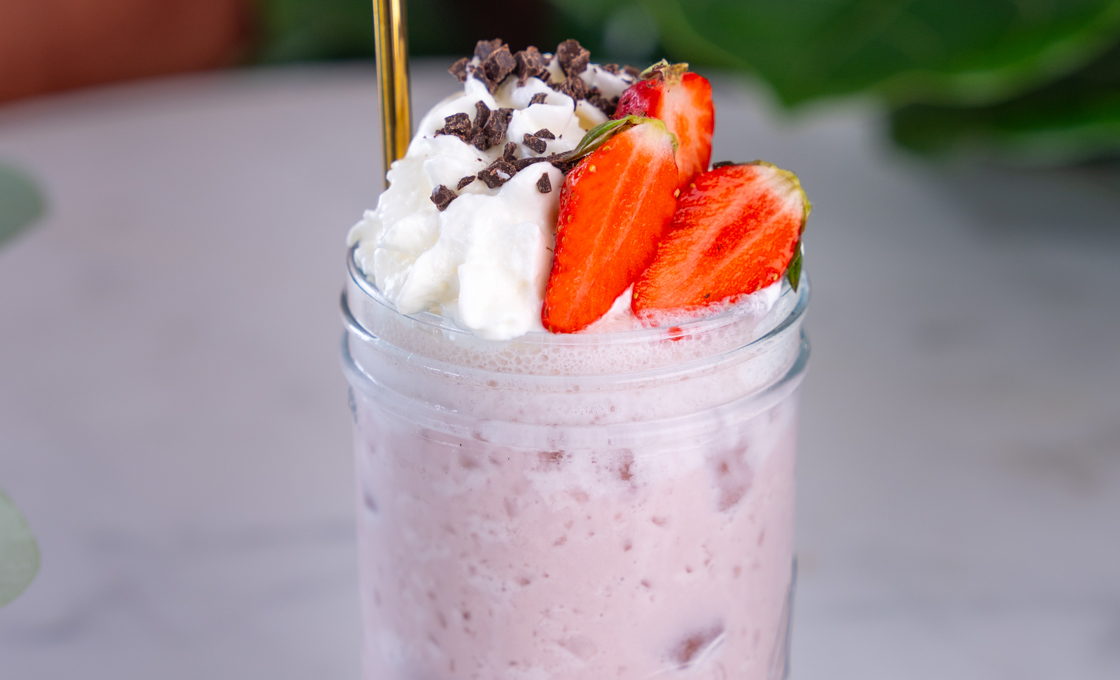 On a marble table top there is a tall milkshake glass, filled with a pink milkshake. The milkshake is topped with cream, strawberry slices and chocolate shavings. 