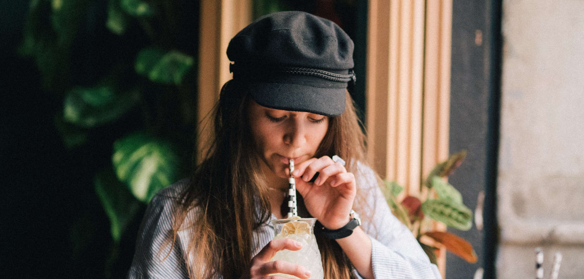 A woman wearing a soft cap drinking a cocktail through a straw