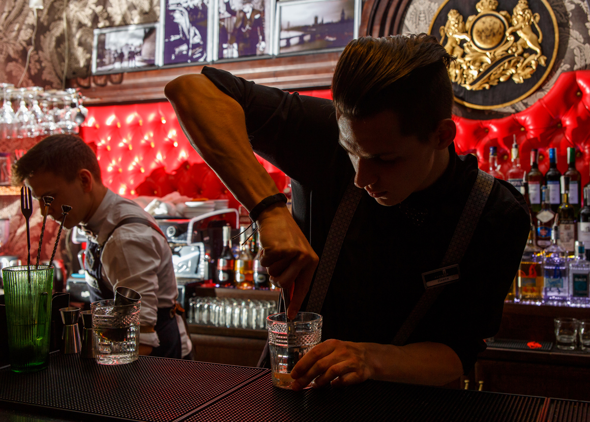 Two bartenders are stood behind a bar. They are both making drinks. One of them is muddling a drink. 