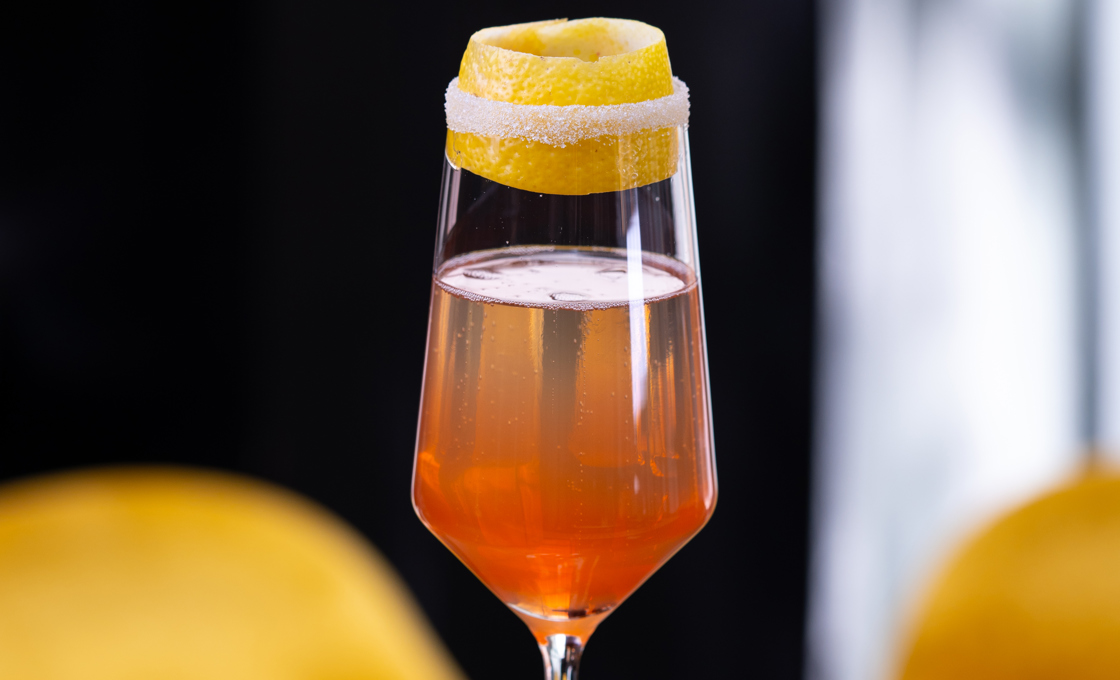 On top of a black bar surface there is a champagne flute glass. The glass is garnished with a slice of lemon peel. In the background there are two yellow bar stalls. 