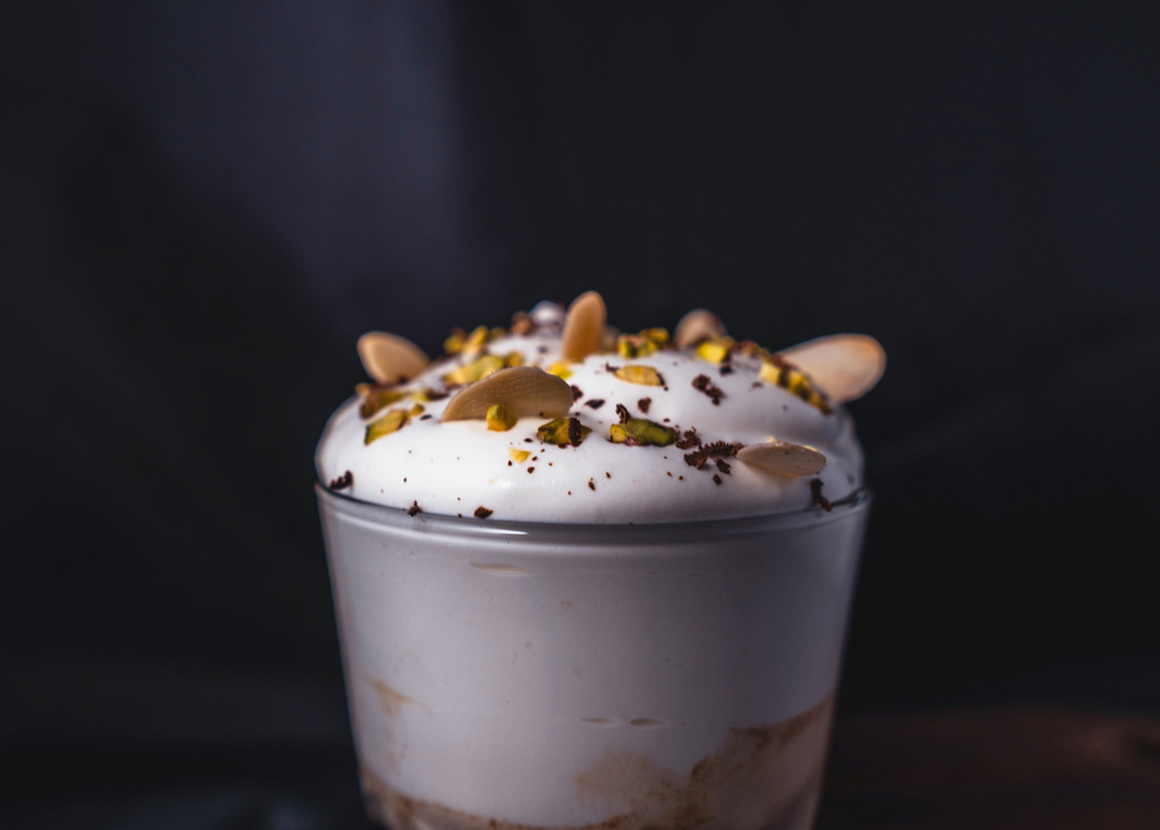 In a tall glass we can see the top of a coffee glass. The top is thick and frothy, and topped with nuts.