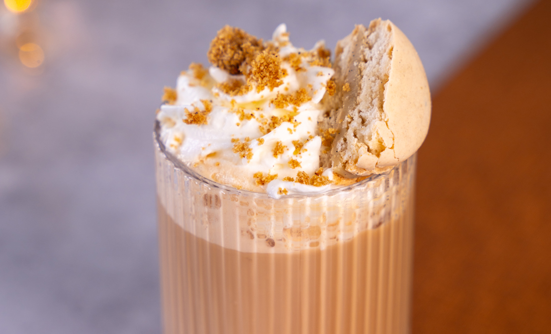 A short tumbler glass placed on a brown matt is atop a marble table. The glass contains a hot chocolate, and is topped with cream and gingerbread crumbs. In the background there are fairy lights. 