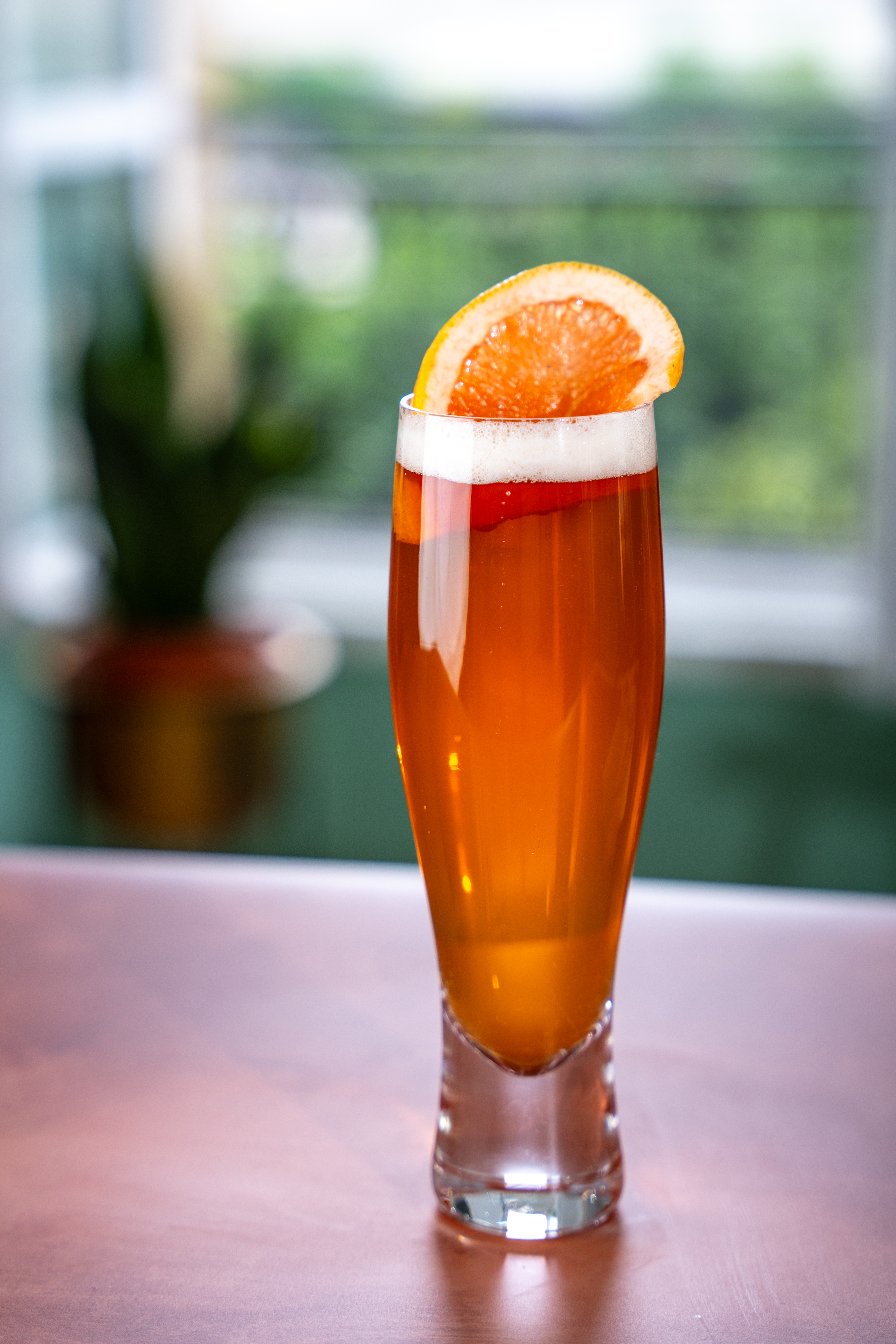 A tall beer glass sits on a polished wooden surface, The glass is filled with an orange coloured drink, and is topped with a slice of pink grapefruit. 