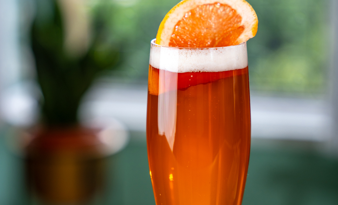 A tall beer glass sits on a polished wooden surface, The glass is filled with an orange coloured drink, and is topped with a slice of pink grapefruit. 
