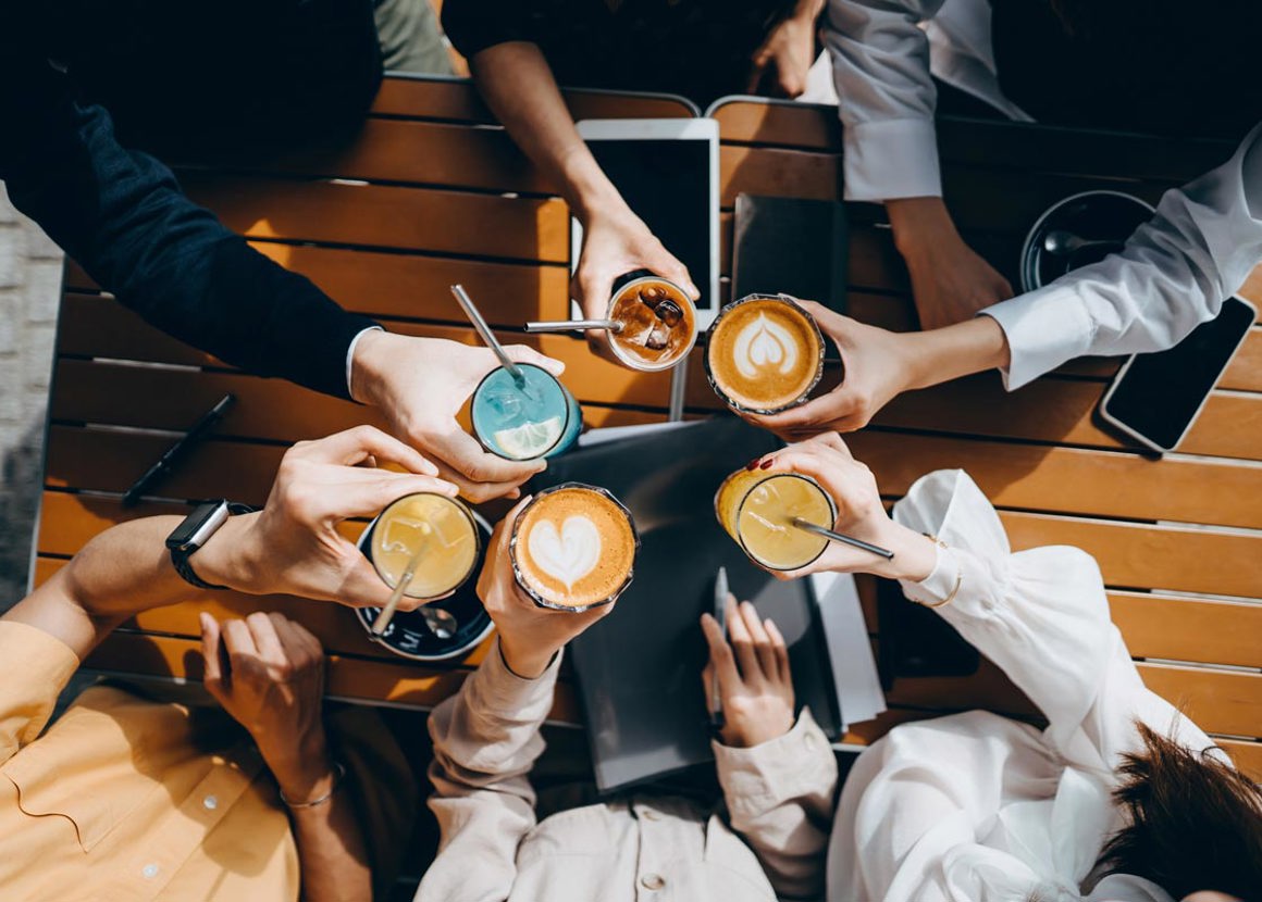 A birds-eye-view of a group of friends sitting outside around a brown wooden table holding cups of coffee and cold sodas. 