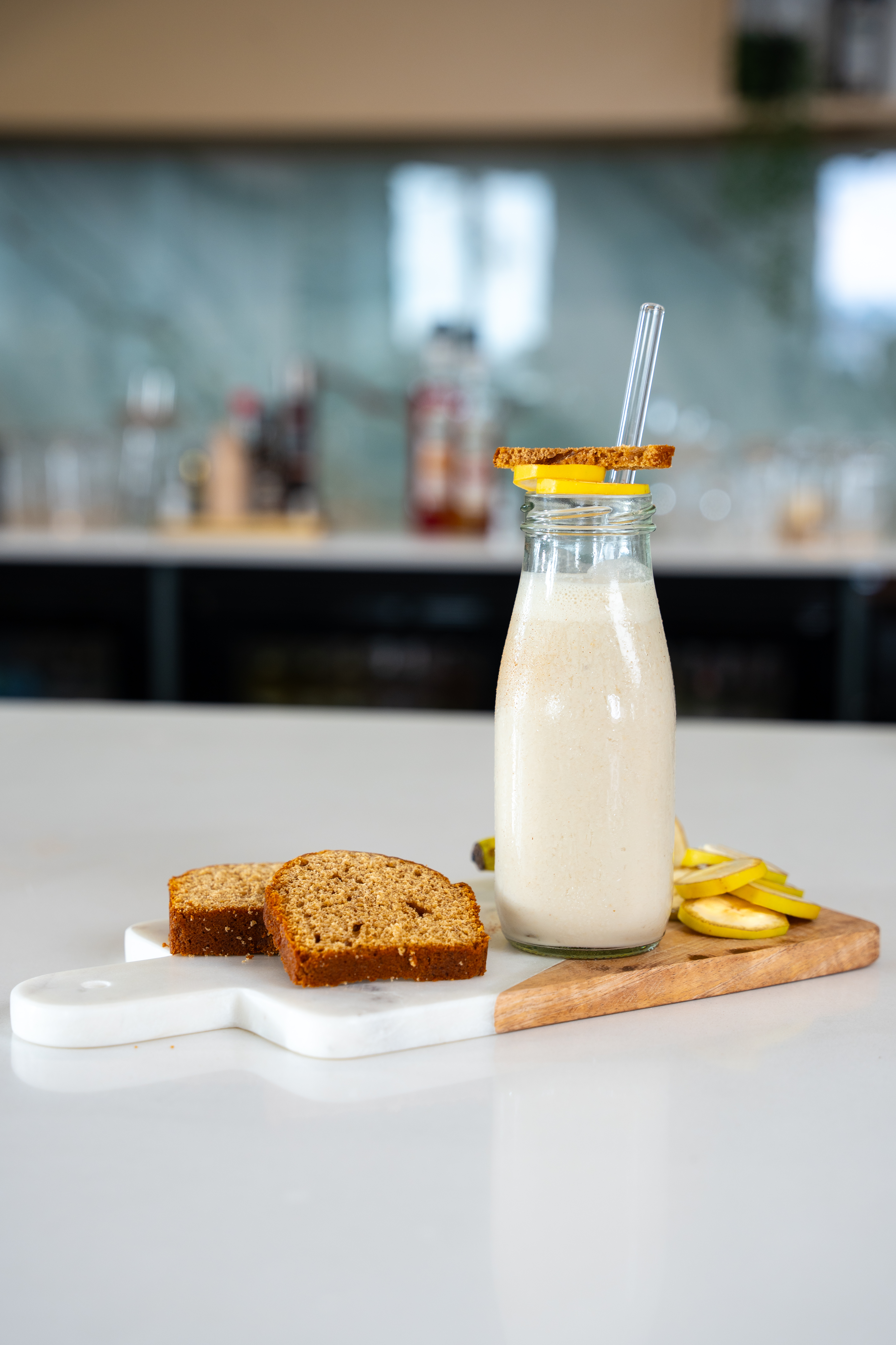On top of a bar surface, placed on a serving board, there is a milk bottle styled glass filled with a cream coloured smoothie. The smoothie is topped with gingerbread and sliced bananas. Also on the serving tray there are slices of gingerbread. 