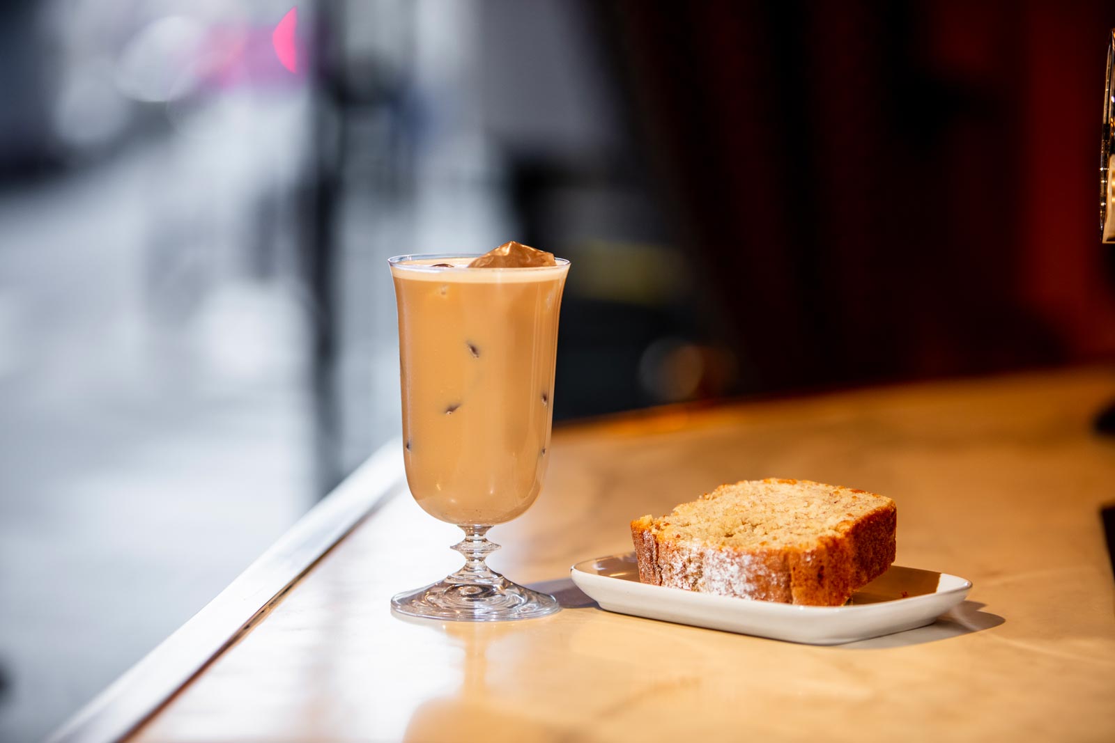 On the surface of a bar there is a slice of banana cake on a small white plate, next to it there is a glass of iced coffee in a tall glass.