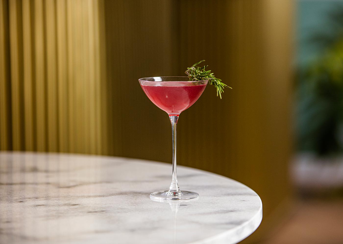 A small coupe glass is placed on the egde of a marble table. The glass contains a red drink and it topped with a sprig of rosemary.