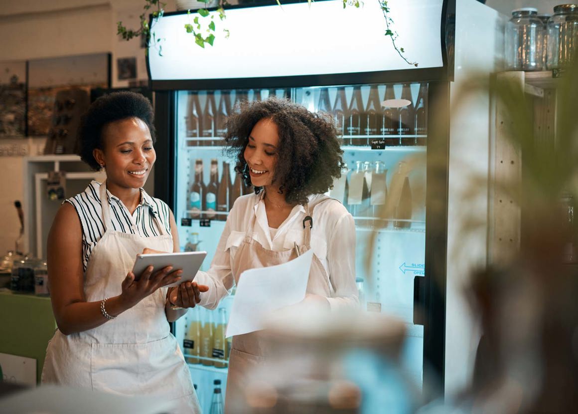 Two females standing in a cafe in front of a drinks fridge. They are looking at an iPad and a sheet of paper as if they are stock taking. 