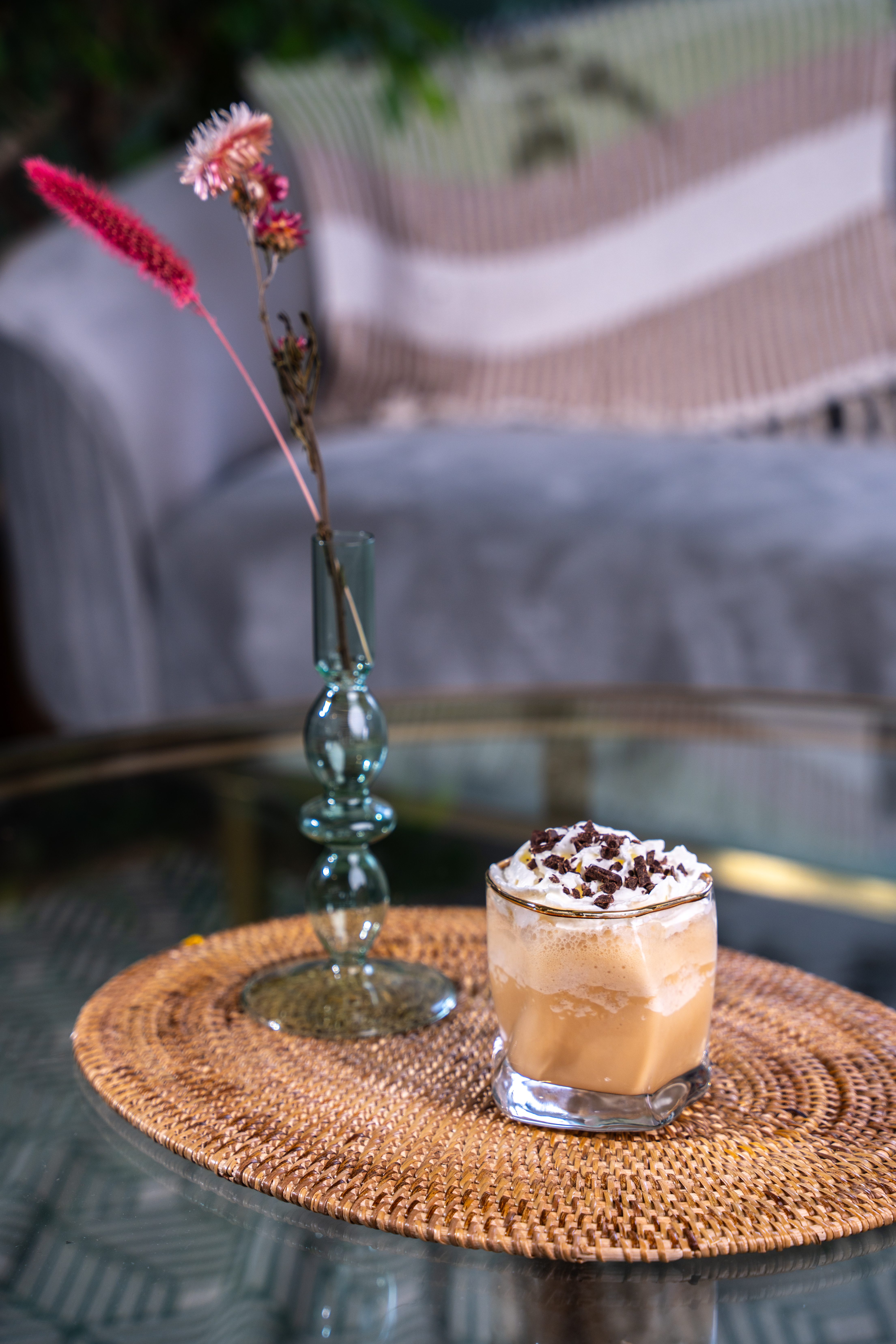 A short tumbler glass is placed on a wicker matt on top of a glass table. The glass contains an iced coffee drink, topped with cream and chocolate shavings. 