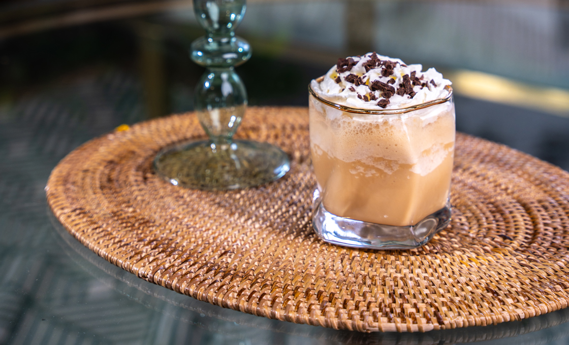 A short tumbler glass is placed on a wicker matt on top of a glass table. The glass contains an iced coffee drink, topped with cream and chocolate shavings. 