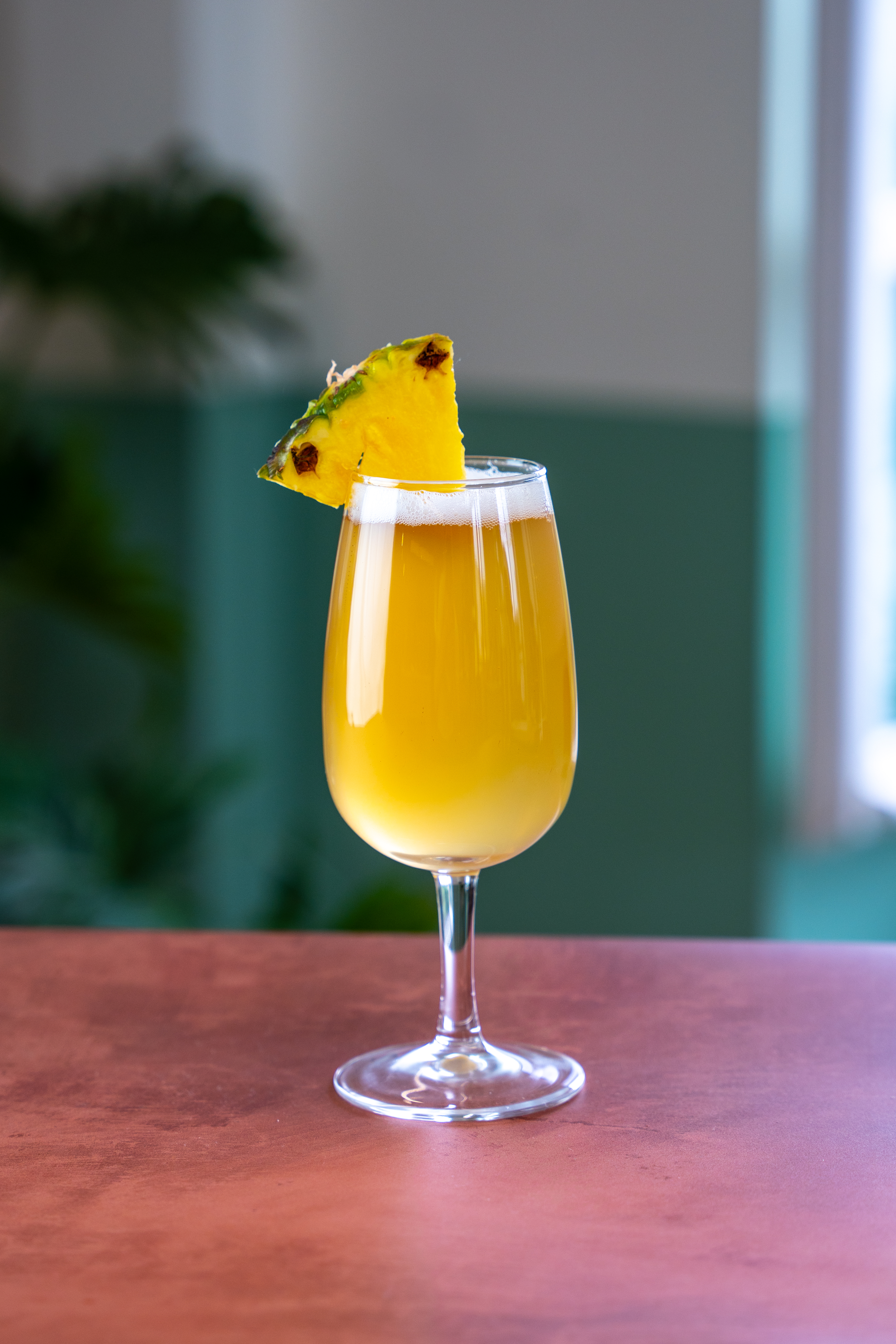 A stemmed beer glass sits on a polished wooden surface. The glass is filled with a yellow coloured drink, and is topped with a pineapple wedge. 