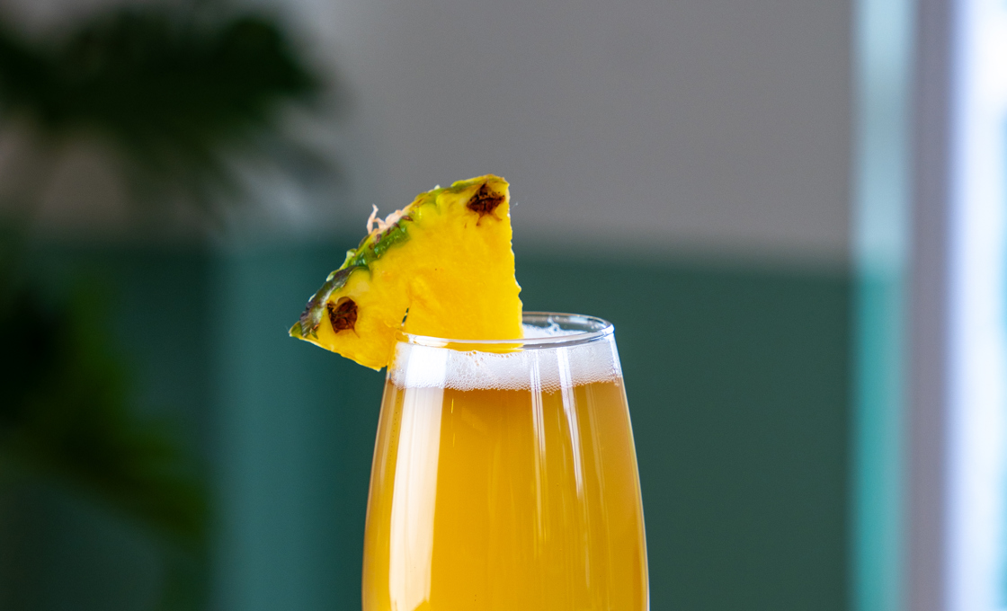 A stemmed beer glass sits on a polished wooden surface. The glass is filled with a yellow coloured drink, and is topped with a pineapple wedge. 