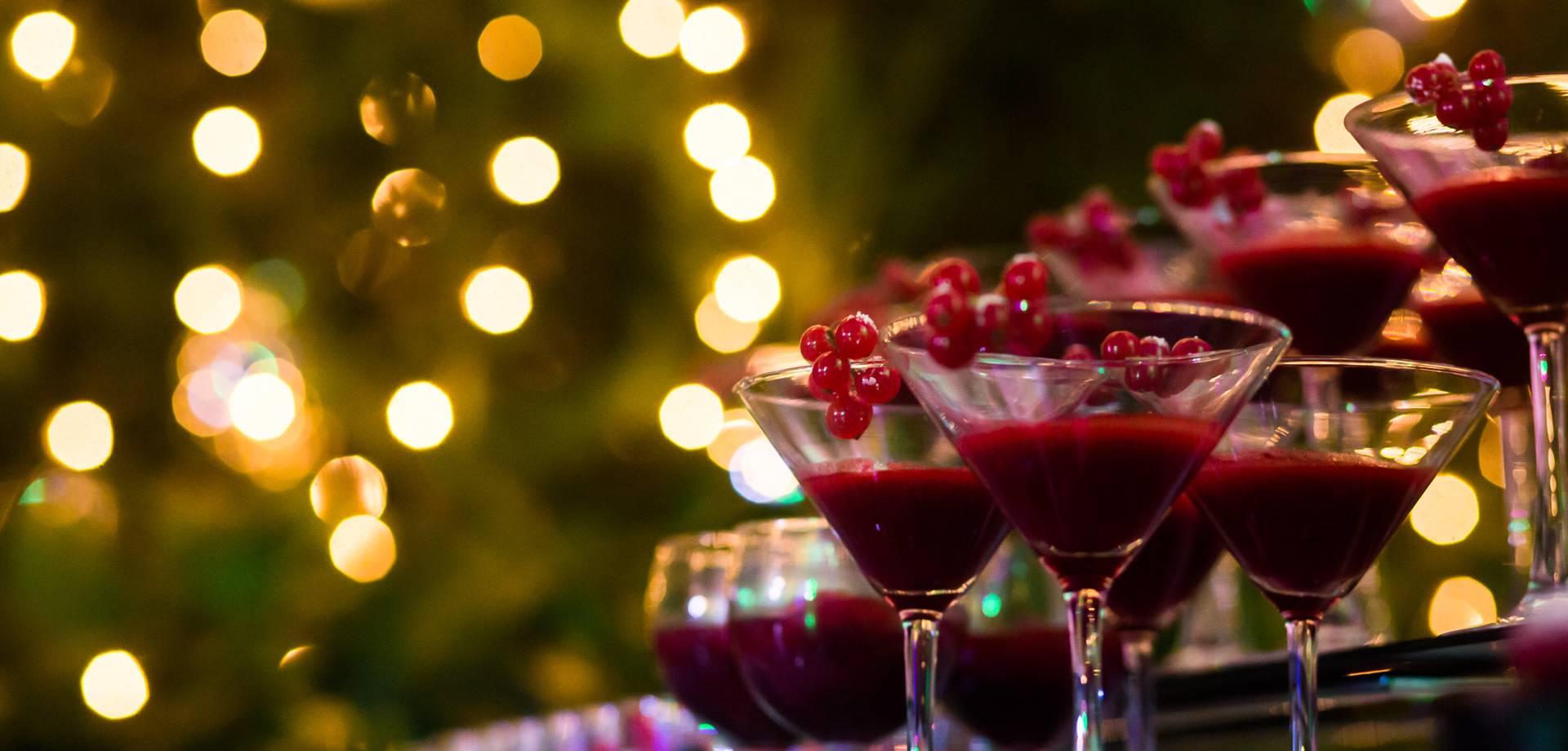 A row of martini glasses containing red drinks are placed along a bar. In the background there are fairy lights.