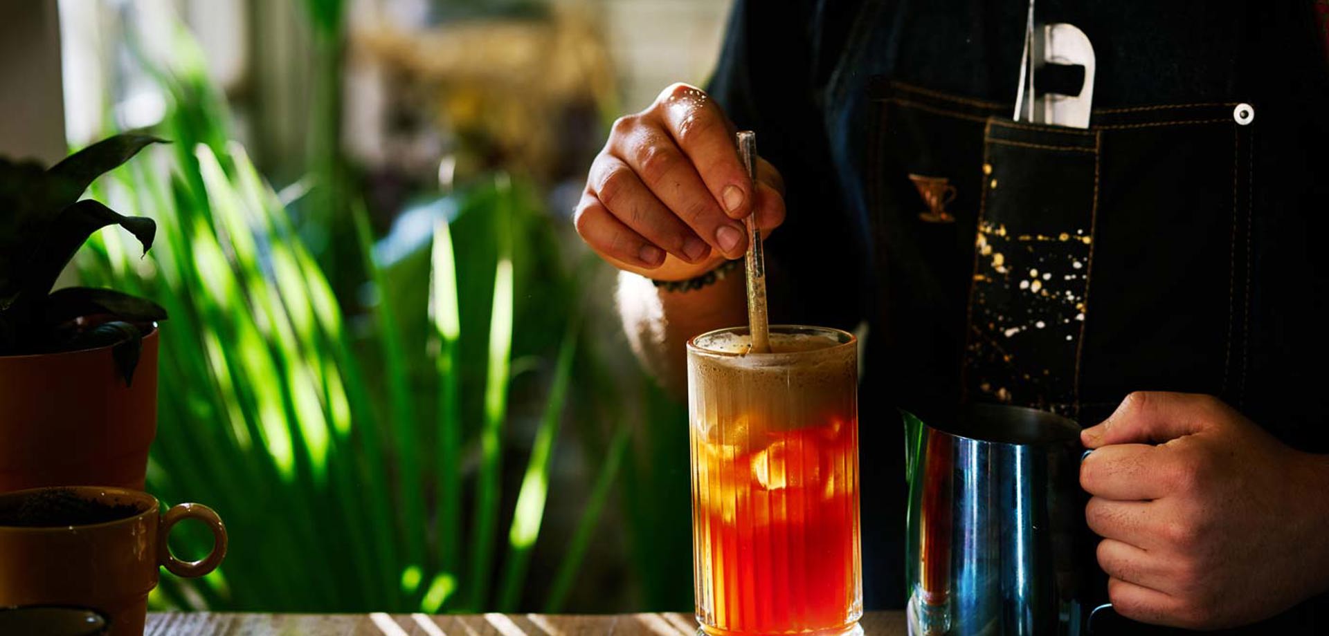 A bartender is mixing a drink in a tall glass. In the background there is a tall plant.