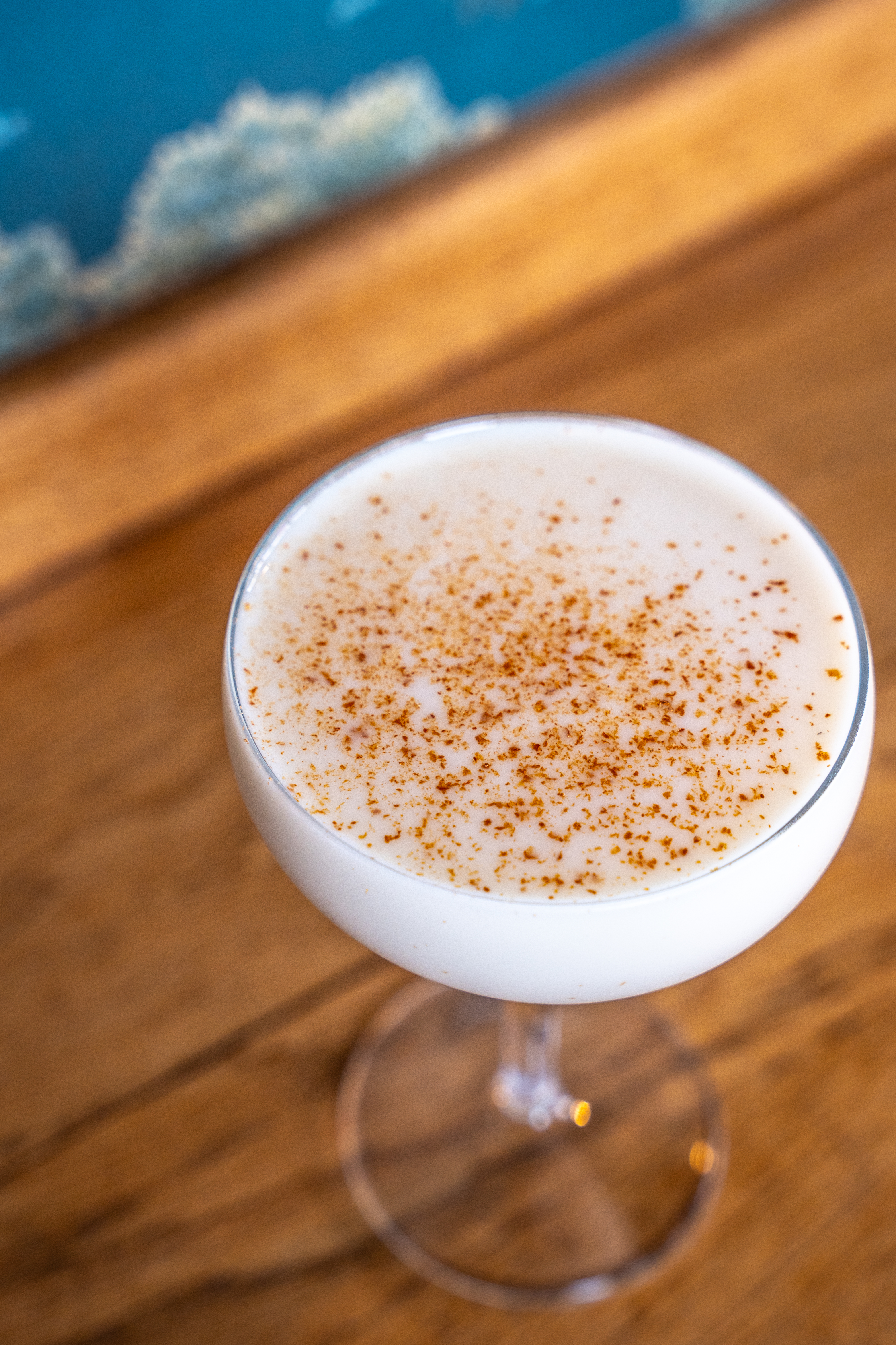 From above we can see a small coupe glass on a wooden surface. The glass is filled with a white drink and is garnished with a sprinkle of macadamia nuts. 