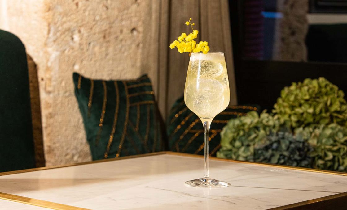 A wine glass is placed on a marble table. The glass contains a clear drink that has been topped with flowers. In the background there are velvet pillows and plants.