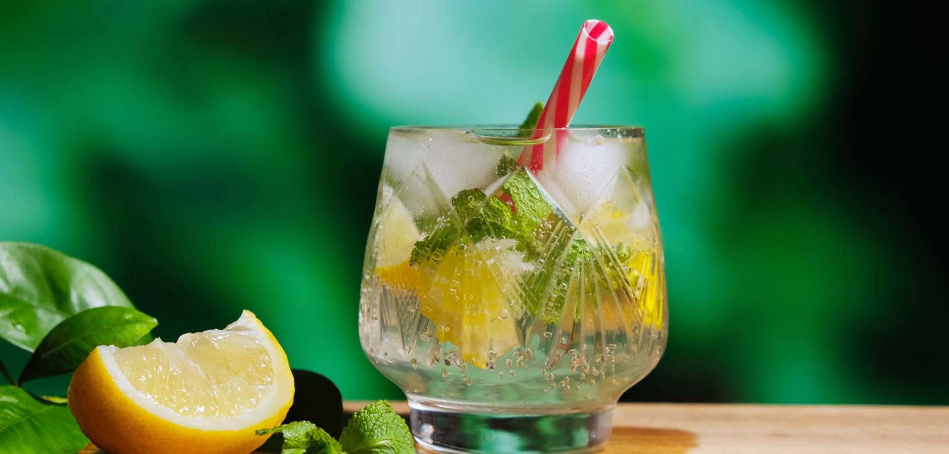 A short glass sits on a wooden table. The glass contains a clear fizzy drink and mint leaves.