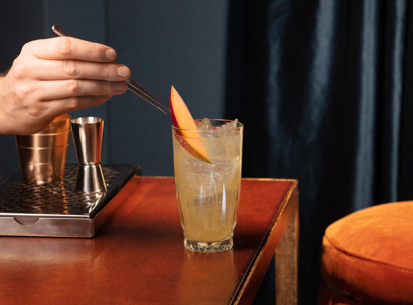 A highball glass is placed at the edge of a wooden bar. The glass contains ice and a pale orange drink. A male hand is holding long tweezers, placing a slice of apple atop the drink as a garnish. In the background there are drinks measures, and an orange bar stool next to the bar.