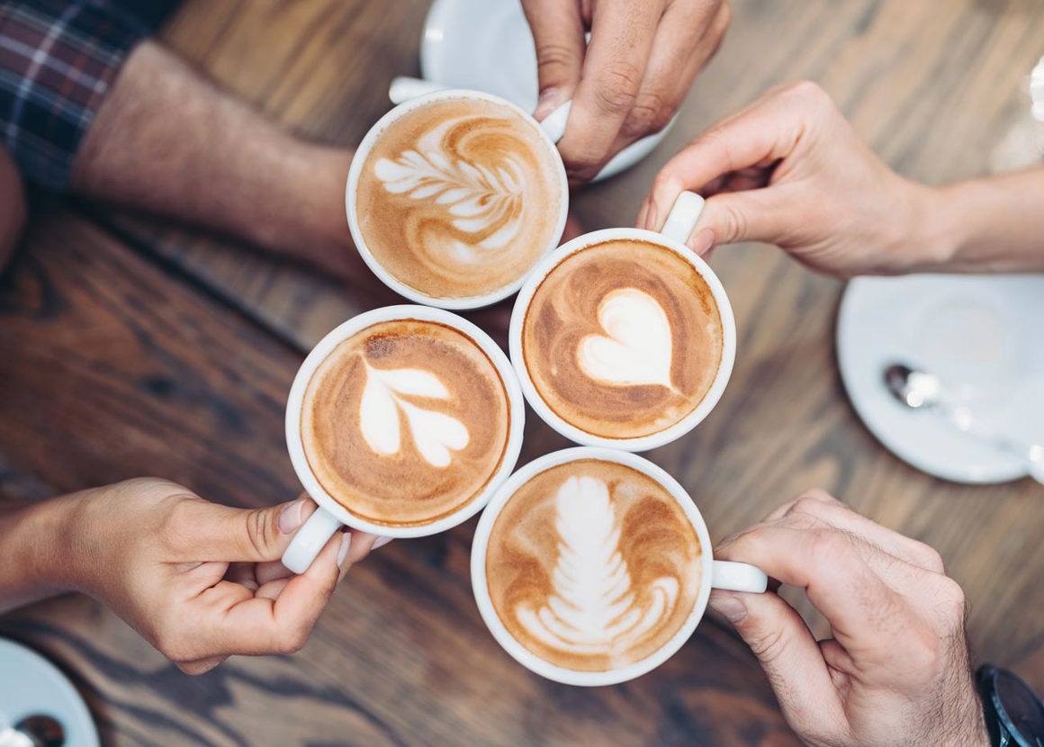 Four people are holding their coffee mugs together. All the drinks have coffee art in the foam, making the shapes of hearts and flowers.