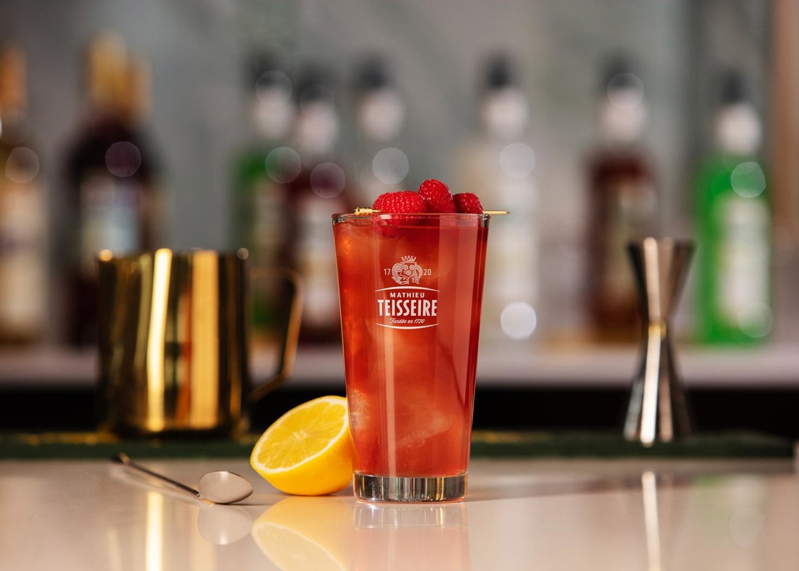 A Mathieu Teisseire glass filled with a red drink is placed on a bar surface. In the background we can see cocktail making equipment.