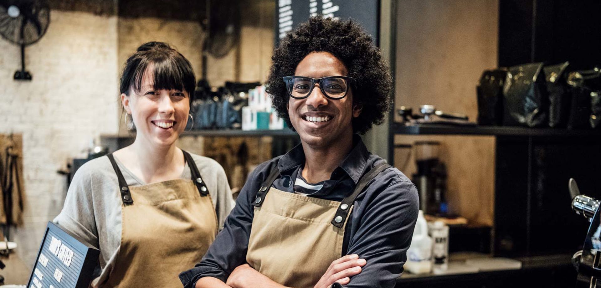 A male and female barista standing in a cafe. They are both wearing brown aprons, smiling at the camera.
