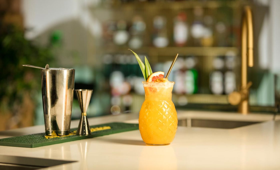A tiki glass containing a yellow punch sits on a bar, garnished with pineapple leaves, a grapefruit wedge and a gold straw. In the background there is cocktail making equipment.
